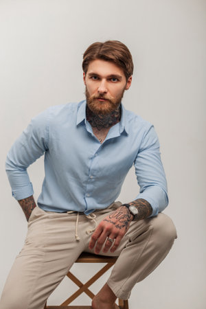 Fashionable young handsome brutal man with beard and hairstyle with tattoos in elegant clothes with shirt sits on wooden chair on a white backgroundの写真素材