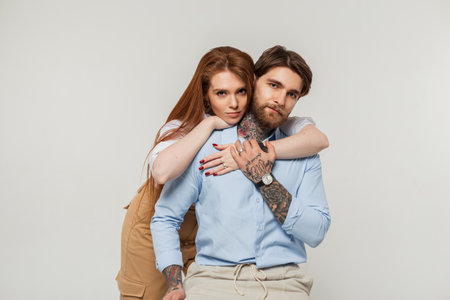 Fashionable beautiful couple, a brutal hipster man with a beard and a tattoos and a beautiful red-haired girl in a shirt on a white background. Handsome guy sits on a chair and a woman hugs himの写真素材