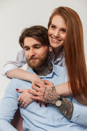 Fashionable beautiful happy couple in the studio. Pretty young beautiful happy red-haired girl with a smile hugs a handsome brutal man with a beard and a tattoosの写真素材