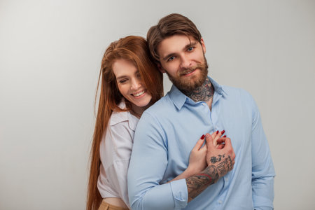 Beautiful fashionable happy couple in the studio. Happy beautiful redhead girl with smile hug handsome brutal hipster man with tattoo and beard in shirtの写真素材