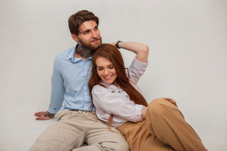 Happy beautiful stylish couple sitting and posing in the studio on a white background. Beautiful fashion red-haired girl with a smile and a handsome fashionable brutal man with a tattoo and a beardの写真素材