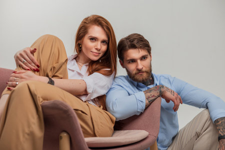 Fashionable beautiful couple in stylish clothes are sitting in a chair on a white background. Beautiful redhead young girl and handsome brutal hipster guyの写真素材
