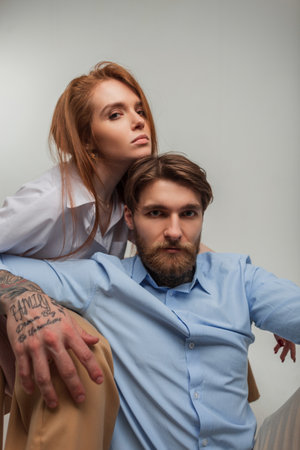 Stylish beautiful young couple in fashionable elegant clothes are sitting on a white background. Beautiful redhead woman model and handsome hipster man with tattoo and beardの写真素材