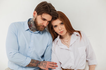 Stylish young couple, beautiful trendy redhead woman and handsome hipster brutal business man with beard and tattoos stand near white backgroundの写真素材