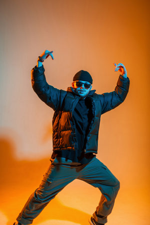 Cool trendy dancer guy with sunglasses in fashion black clothes with a black down jacket and hat dances in a creative studio with yellow and blue lightの写真素材
