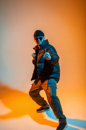 Fashionable happy professional dancer man with hat and sunglasses in fashion black down jacket posing in studio with creative orange and neon lightの写真素材