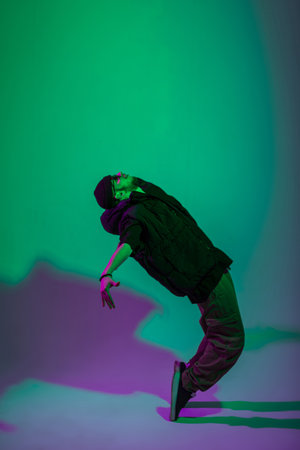 Fashionable handsome professional hip hop dancer man with a hat in a vest, jeans and sneakers dances in motion in a creative studio with green and purple lightの写真素材