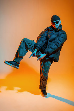 Fashion handsome young hip hop dancer guy with sunglasses and a hat in fashionable clothes with a down jacket and sneakers is dancing in a color studio with orange and neon lightの写真素材