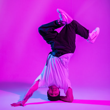 Fashionable handsome hipster b-boy dancer guy in stylish clothes dances on one arm in a creative...
