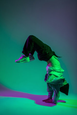 Fashion handsome hip hop dancer man b-boy in trendy denim clothes dancing in motion and standing on his hands in a creative color studio with green and purple lightの写真素材