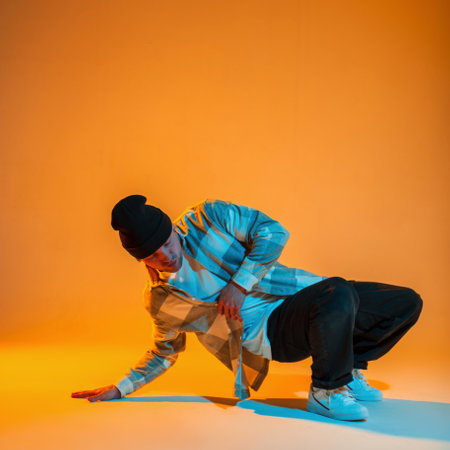 Stylish beautiful professional dancer in trendy fashion clothes with a shirt, hat, jeans and white sneakers dances on the floor in a creative color studio with orange and neon lightsの写真素材