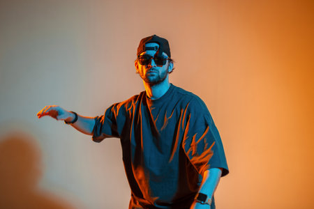 Stylish handsome cool professional dancer man with a cap and sunglasses in trendy fashion clothes is dancing in a creative colorful studio with warm orange and cold neon lightの写真素材