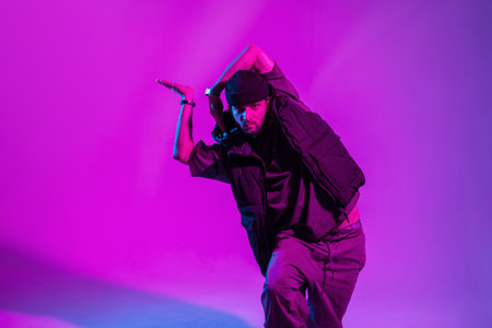 Fashionable professional male dancer in trendy fashion clothes with a hat and vest dancing in a creative color studio with pink neon lightsの写真素材