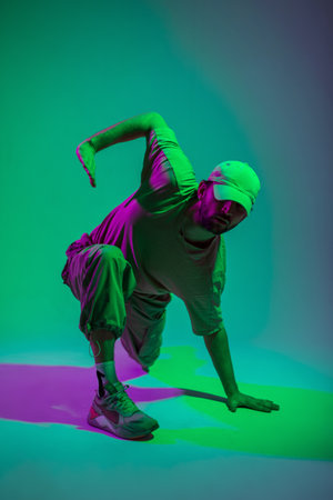 Cool trendy professional hip hop dancer man with cap in fashion clothes dancing in a colorful studio with green and pink lightsの写真素材