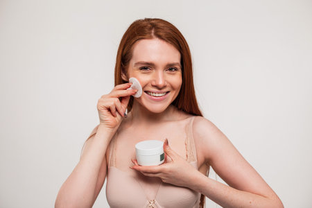 Beautiful happy young fresh redhead with smile girl is holding a cream jar and wiping her face with a cotton pad in the studio. beauty and skin careの写真素材