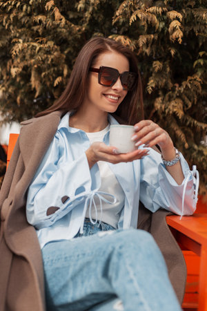Beautiful happy girl with a smile with sunglasses in fashionable casual clothes drinks tea and enjoys the spring sunny weatherの写真素材