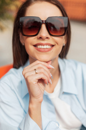 Happy street portrait of a beautiful cheerful girl with a smile with elegant vintage sunglasses in fashion casual clothes sits and rests on a sunny spring dayの写真素材