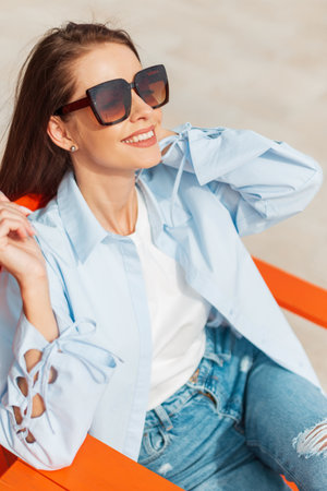 Elegant beautiful young happy girl with a smile with cool vintage sunglasses in fashionable casual clothes with a shirt and jeans sits and enjoys a warm sunny spring dayの写真素材