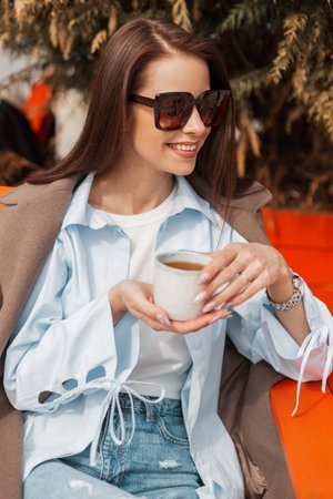 Happy beautiful young woman with a smile with sunglasses in a fashionable coat and shirt holds a cup and drinks tea outdoors on a sunny dayの写真素材