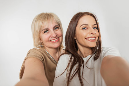 Beautiful young happy girl daughter and pretty elderly cheerful mother woman with a smile make a selfie photo on a white backgroundの写真素材