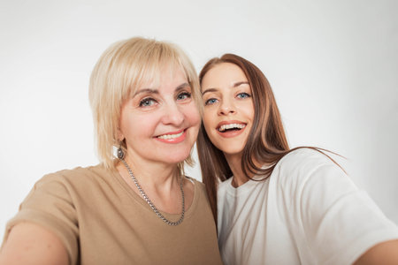 Beautiful happy young daughter woman with a smile and a cheerful elderly pretty mother take a selfie photo together on a white background in the studioの写真素材