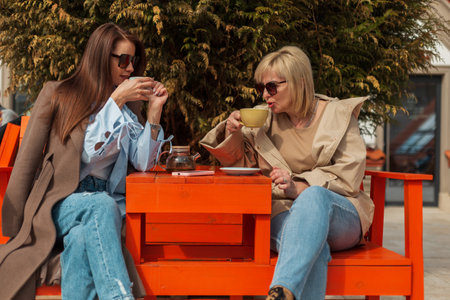 Fashionable beautiful mother and daughter in stylish casual clothes sit in a cafe on the street and drink tea and coffee on a spring sunny dayの写真素材