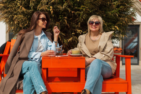 Cheerful beautiful elderly mother and happy daughter with smile with sunglasses in trendy casual clothes sit outside in a cafe and drink tea with coffee on a sunny dayの写真素材