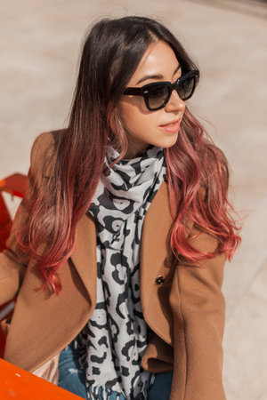 Fashion beautiful female model with pink hair with vintage sunglasses in a trendy stylish coat and scarf sits on the street on a sunny spring dayの写真素材