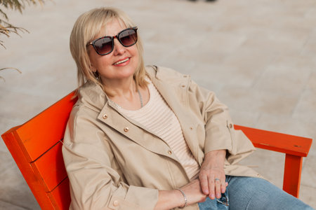 Fashionable pretty elderly happy woman with a smile with sunglasses in trendy stylish clothes with a beige jacket and sweater sits and rests outdoors on a sunny warm spring dayの写真素材