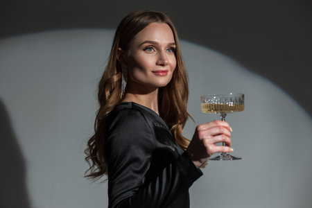 Elegant beautiful chic lady with an earring in a trendy fashion black dress with a vintage champagne glass celebrating an event on a dark background with circular light. pretty womanの写真素材