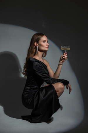 Elegant beautiful young woman model with diamond earrings in a fashionable evening black dress with a vintage champagne glass sits on a dark background in the studio. Pretty lady celebrate an eventの写真素材
