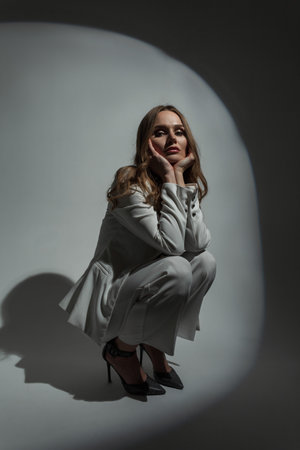 Fashion beautiful glamorous woman in a white suit with a blazer and heels sits and poses on a dark background in the studioの写真素材