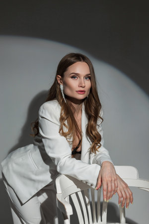 Beautiful fashionable lady in a white stylish suit stands near a white chair on a dark background in the studio at a partyの写真素材