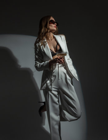 Fashion beautiful chic woman with cool sunglasses in a stylish white suit with a blazer and a bra with a glass of champagne on a dark background with a round light at a party. lady celebratingの写真素材