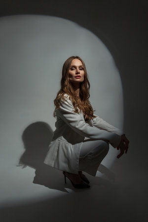 Fashionable beautiful stylish woman model in fashion white suit with blazer sitting and posing in studio on dark backgroundの写真素材