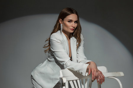 Stylish elegant business lady with a diamond earring in a fashionable white suit with a bra posing near a white chair on a dark background in the studio. Pretty chic woman at partyの写真素材