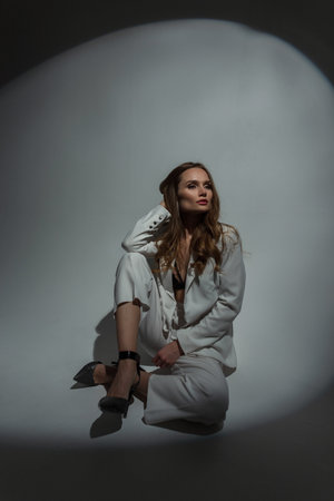 Stylish beautiful elegant fashionable woman model in a fashion white suit with heels shoes sits on the floor in the studioの写真素材
