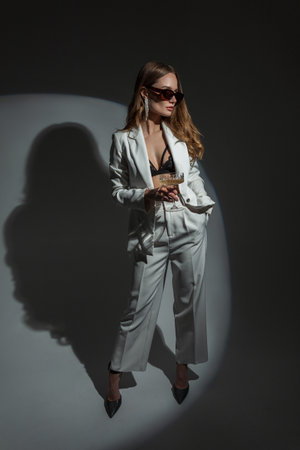 Fashionable beautiful woman in a stylish elegant white suit with a blazer and a bra with heels holds a glass of champagne and poses in the studio on a dark background with circular light. Pretty ladyの写真素材