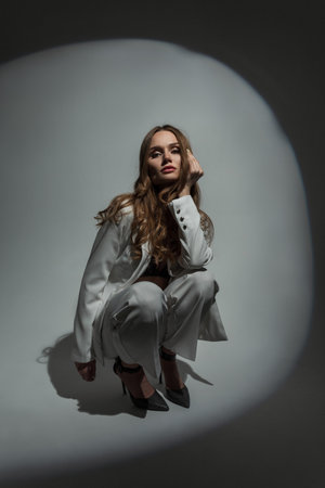Fashionable beautiful stylish chic woman model in fashion white suit sitting and posing in studio on dark background at partyの写真素材