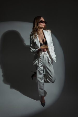 Elegantly pretty woman with sunglasses in white stylish suit with blazer, bra and heels shoes in studio with circle light at party. Chic lady with vintage glass of champagneの写真素材