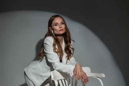 Beautiful stylish successful business girl in an elegant white suit with blazer stands and poses near a white chair in the studio. Pretty chic lady at partyの写真素材