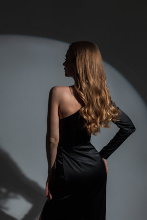 Beautiful elegant woman model with curly brunet hair in fashionable black dress, back view. Chic lady on dark background in studio with circular lightの写真素材