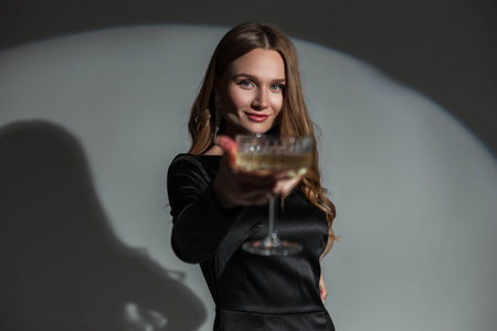 Beautiful happy stylish chic woman with a smile in a fashionable black elegant dress raises a glass of champagne on a dark background in the studio. Pretty lady at partyの写真素材