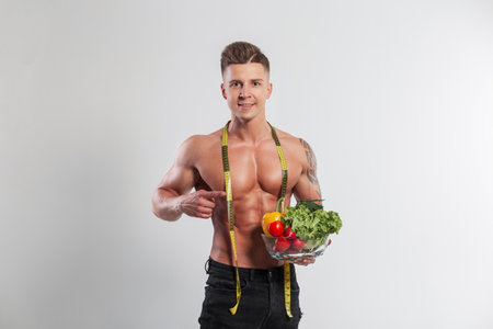 Athletic handsome happy man with a muscular body and a centimeter tape holds and points to a glass bowl with vegetables on a white background in the studio. Healthy food and diet conceptの写真素材