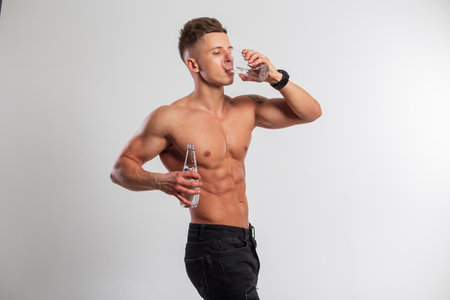 Handsome muscular athletic man with a naked torso holds a bottle of water and drinks water from a glass on a white background. water balance and healthの写真素材