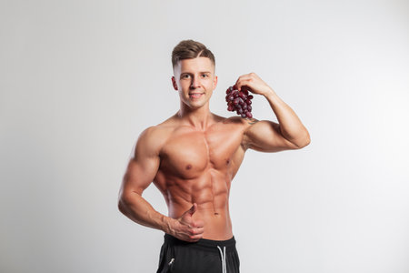 Handsome cool strong healthy athletic man with muscular body holding grapes and showing like on white background. Diet and natural vitaminsの写真素材