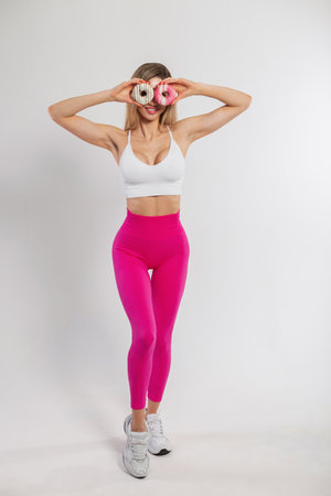 Funny beautiful young fitness lady in fashionable sportswear holds donuts near her eyes and poses on a white backgroundの写真素材