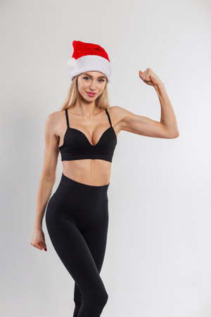 Happy beautiful fitness blonde girl with a slim body with a black bra and red santa claus cap shows biceps on a white background. New Year and sport conceptの写真素材