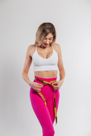 Happy beautiful young fitness lady with a slim body measures her waist with a centimeter tape on a white backgroundの写真素材