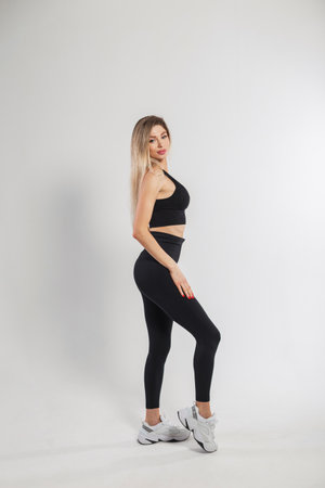 Beautiful young blonde fitness girl in fashionable black sportswear with sneakers, leggings and a top on a white background in the studioの写真素材
